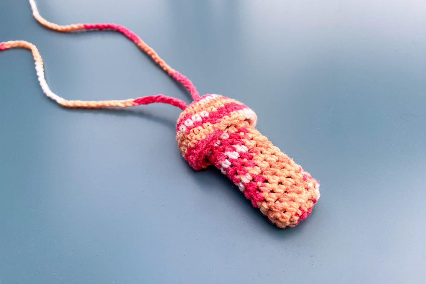 Ein gehäkelter Feuerzeughalter zum Umhängen inin Pilzoptik in bunten Farben liegt auf blauem Hintergrund.