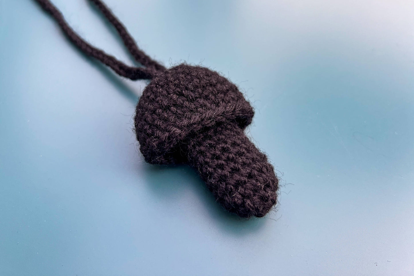 Ein gehäkelter Feuerzeughalter zum Umhängen in Pilzoptik in schwarz liegt auf blauem Hintergrund.