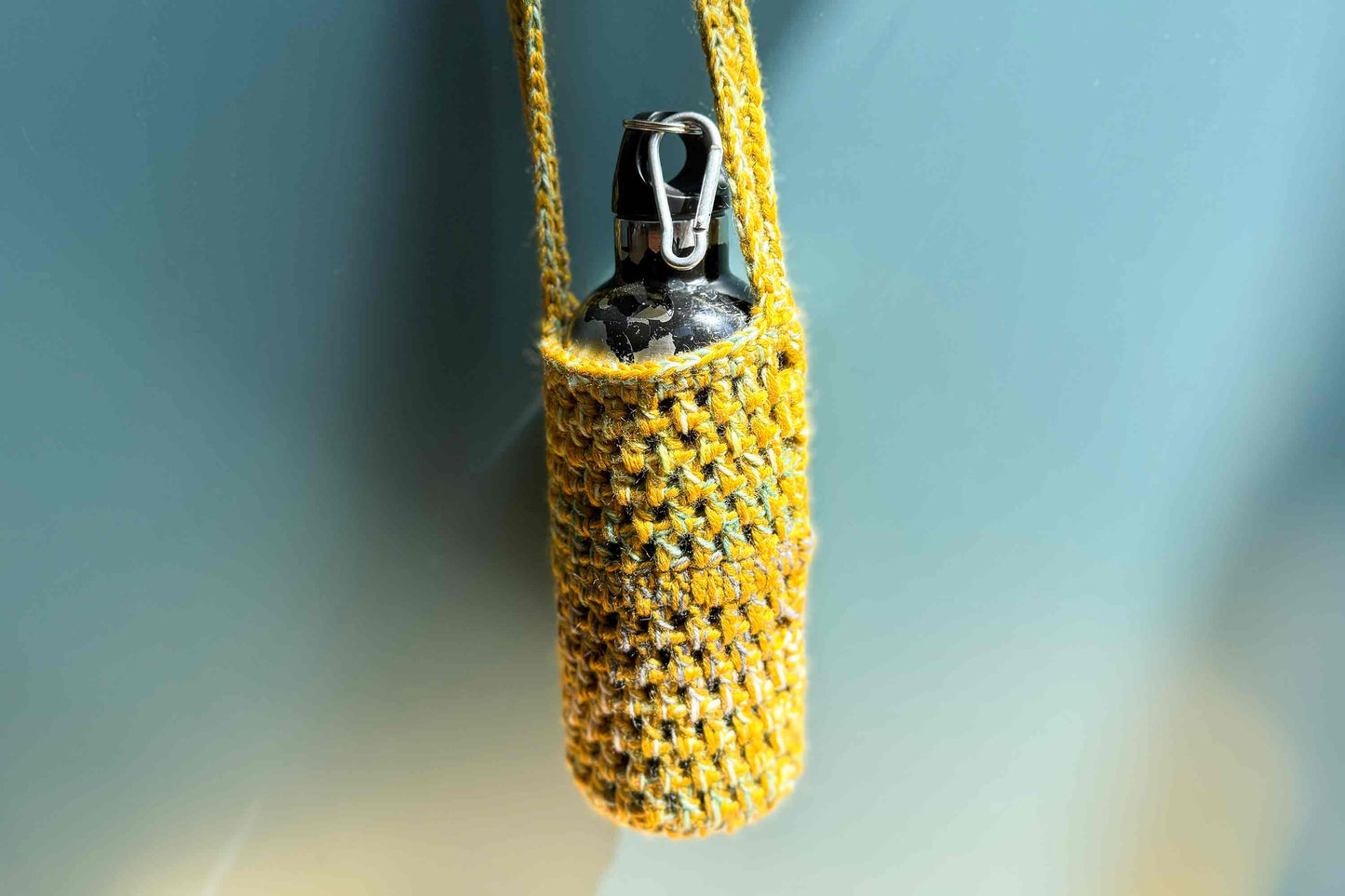 Hängender gehäkelte Flaschenhalter zum Umhängen aus gelber, geretteter Wolle vor blauem Hintergrund