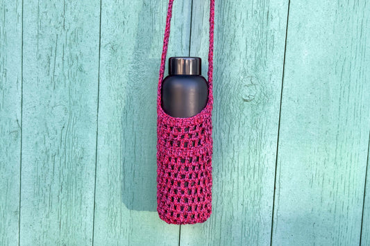 Gehäkelter, Flaschenhalter CHRISTA von Jeff Every Piece A Friend vor blauem Hintergrund. Der Flaschenhalter hat die Farbe pink oder magenta gemischt.