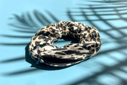 Ein Scrunchie mit Reißverschluss und klassichem braun-weiß-grauem Leopardenmuster liegt auf einem blauen Tisch. Der Reißverschluss ist geöffnet.