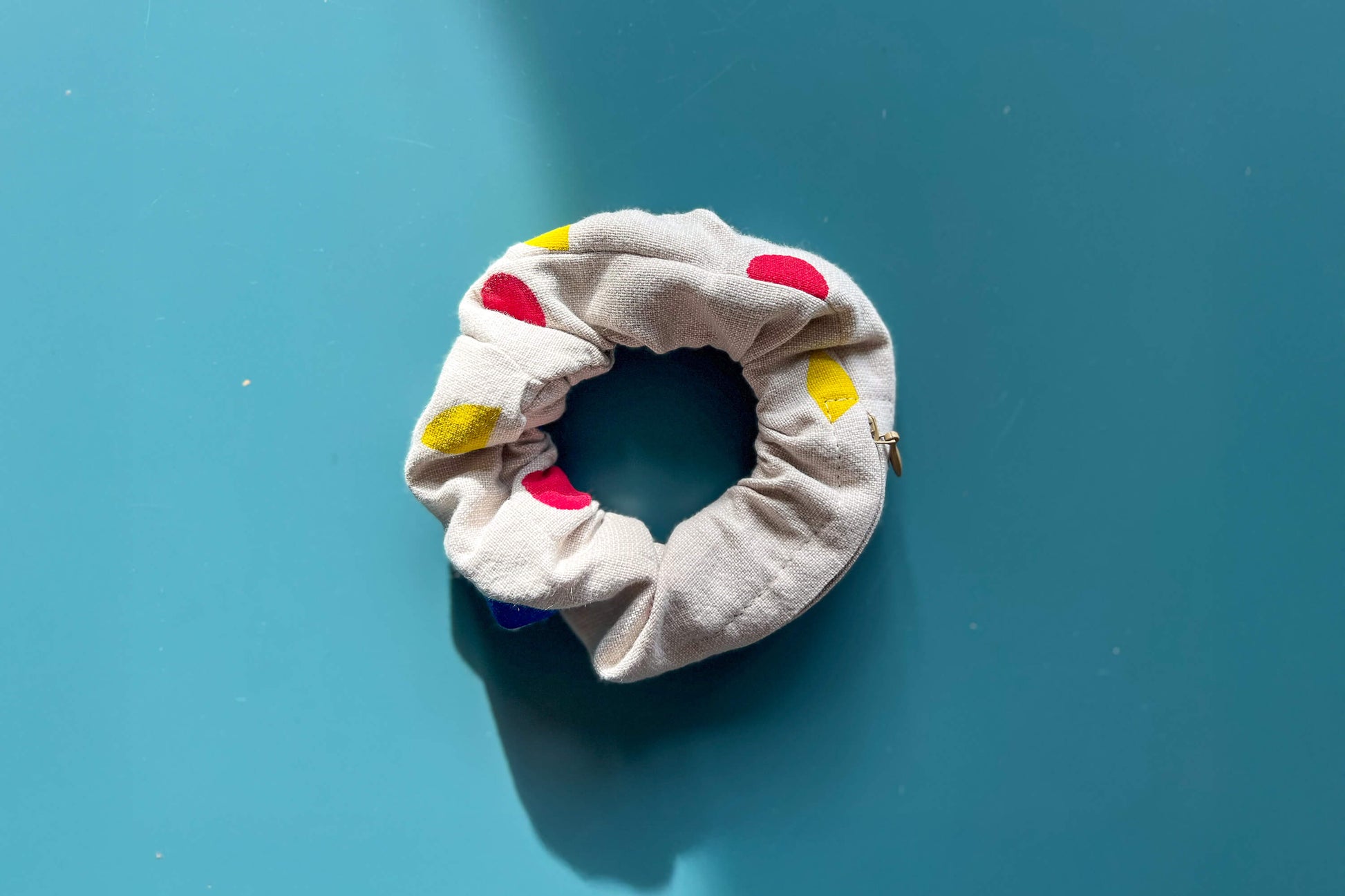 Ansicht von oben: ein beiger Scrunchie mit beigen Reißverschluss, gemustert mit geometrischen, roten, blauen und gelben Flächen liegt auf einem blauen Tisch.