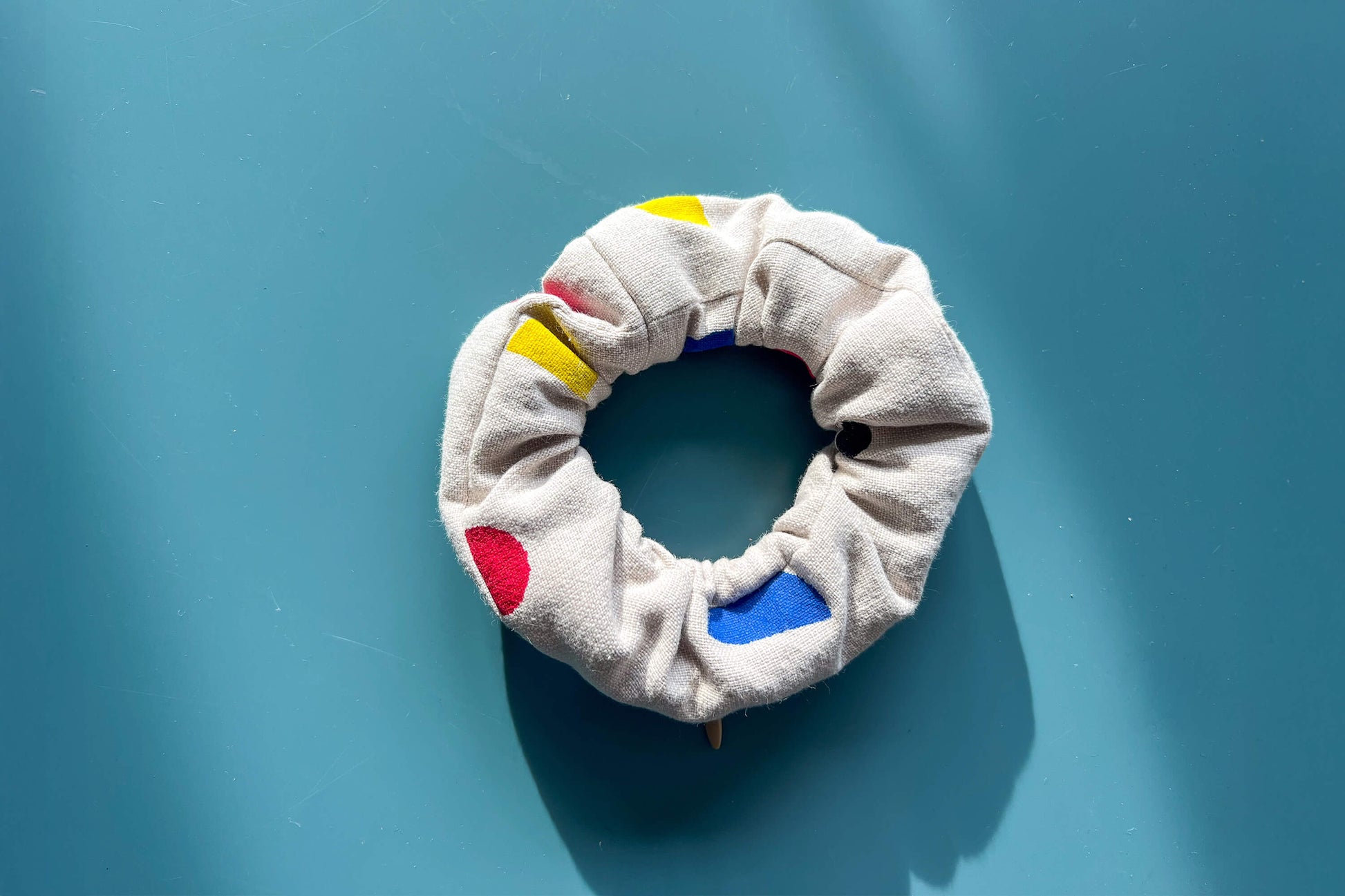 Ansicht von oben: ein beiger Scrunchie mit beigen Reißverschluss, gemustert mit geometrischen, roten, blauen und gelben Flächen liegt auf einem blauen Tisch. Der Reißverschluss ist geschlossen.
