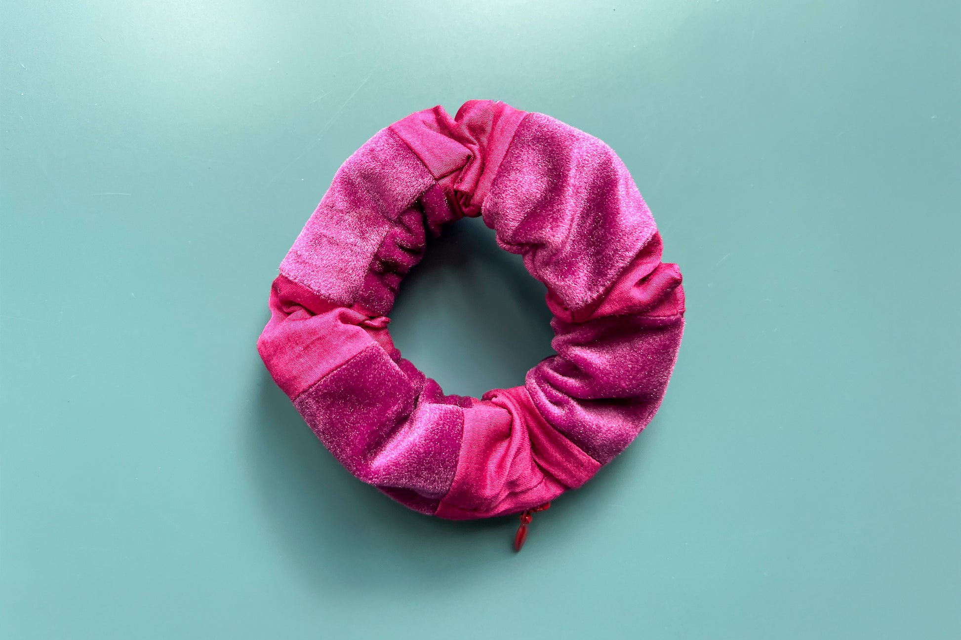 Ansicht von oben: Ein magenta-lila Scrunchie mit Reißverschluss und liegt auf einem blauen Tisch. Der Schrunchie besteht aus magenta Baumwoll- und lila Samstoff. Er hat einen Reißverschluss, dieser ist Reißverschluss geschlossen.