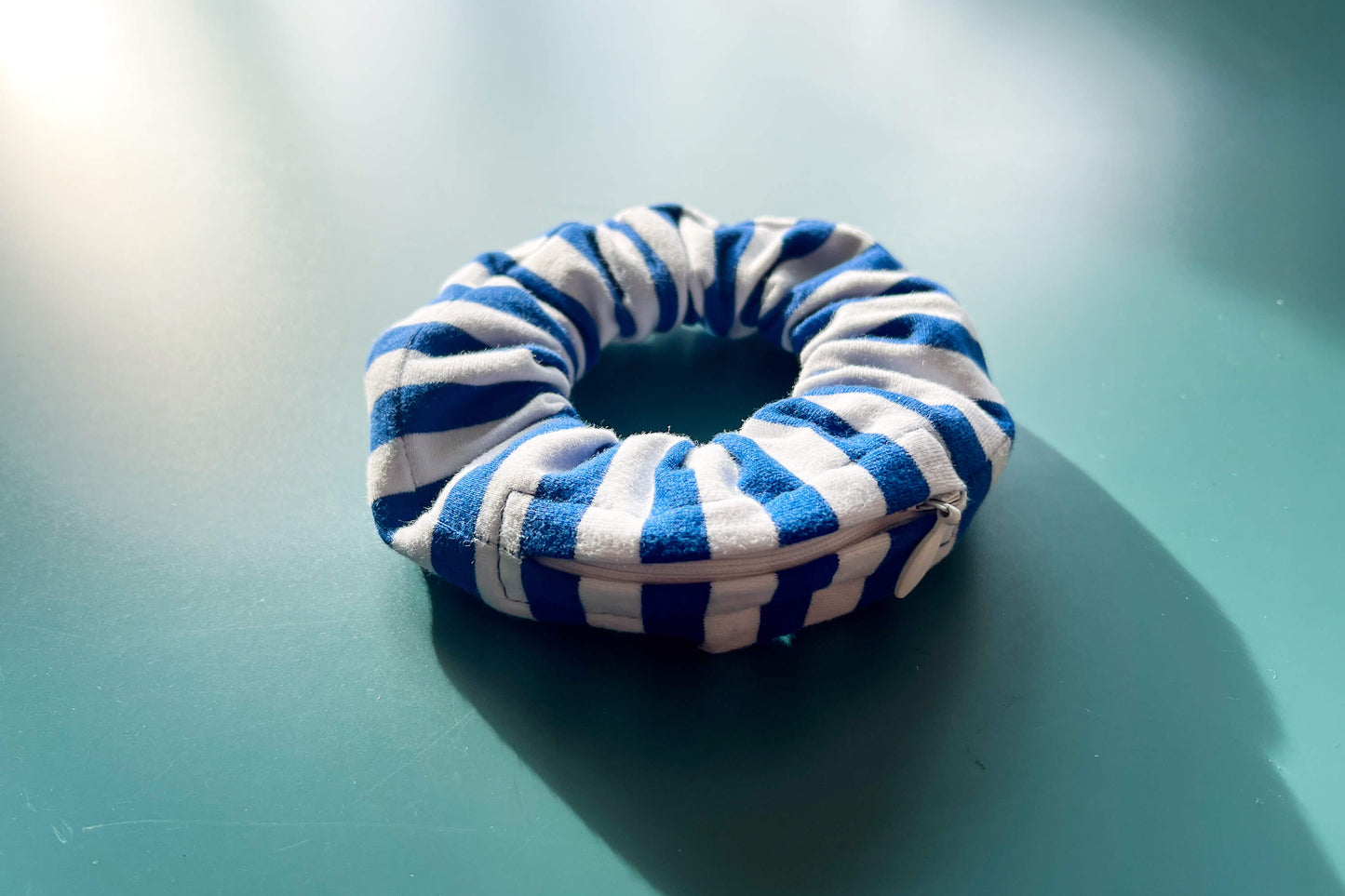 Anisicht von schräg oben: ein Scrunchie mit Reißverschluss und einem Muster in hellblau und weiß liegt auf einem blauen Tisch. Der Reißverschluss ist geschlossen und hat die Farbe weiß.