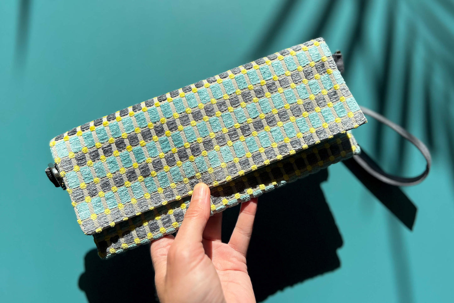 Hand hält die kleinen Umhängetasche DANA aus upcycled Polsterstoff in den Farben blau, gelb und beige mit silbernen Verschlüssen vor einem blauen Hintergrund in die Kamera.