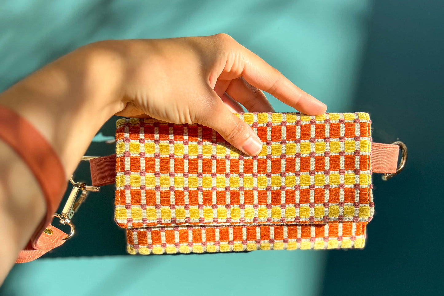 Gesamtansicht: eine Hand hält die Handtasche DANA in der Farbe gelb-orange-beige vor einem blauen Hintergrund ins Bild.