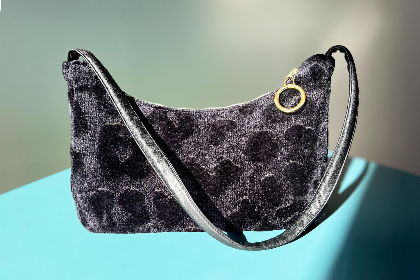 Gesamtansicht: Schultertasche im Vintagestil vor blau-grauem Hintergrund. Die Tasche ist schwarz gemustert und aus Samt und hat einen goldenen Reißverschluss. Die Tasche wurde aus upcycled Materialien von Jeff Every Piece A Friend hergestellt