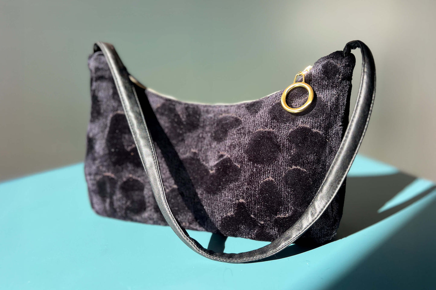 Gesamtansicht: Schultertasche im Vintagestil vor blau-grauem Hintergrund. Die Tasche ist schwarz gemustert und aus Samt und hat einen goldenen Reißverschluss. Die Tasche wurde aus upcycled Materialien von Jeff Every Piece A Friend hergestellt