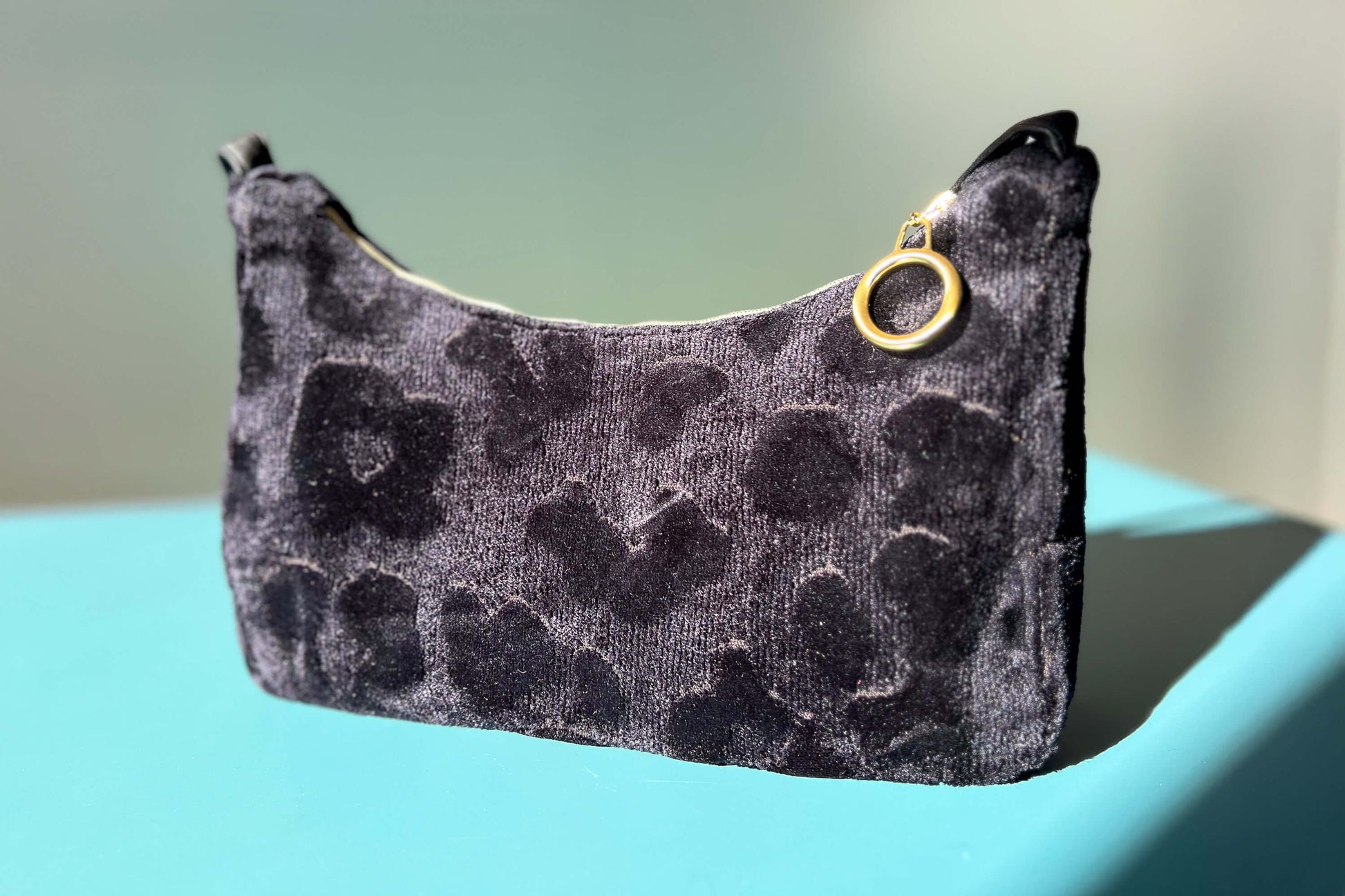 Gesamtansicht: Schultertasche im Vintagestil vor blau-grauem Hintergrund. Die Tasche ist schwarz gemustert und aus Samt und hat einen goldenen Reißverschluss. Die Tasche wurde aus upcycled Materialien von Jeff Every Piece A Friend hergestellt