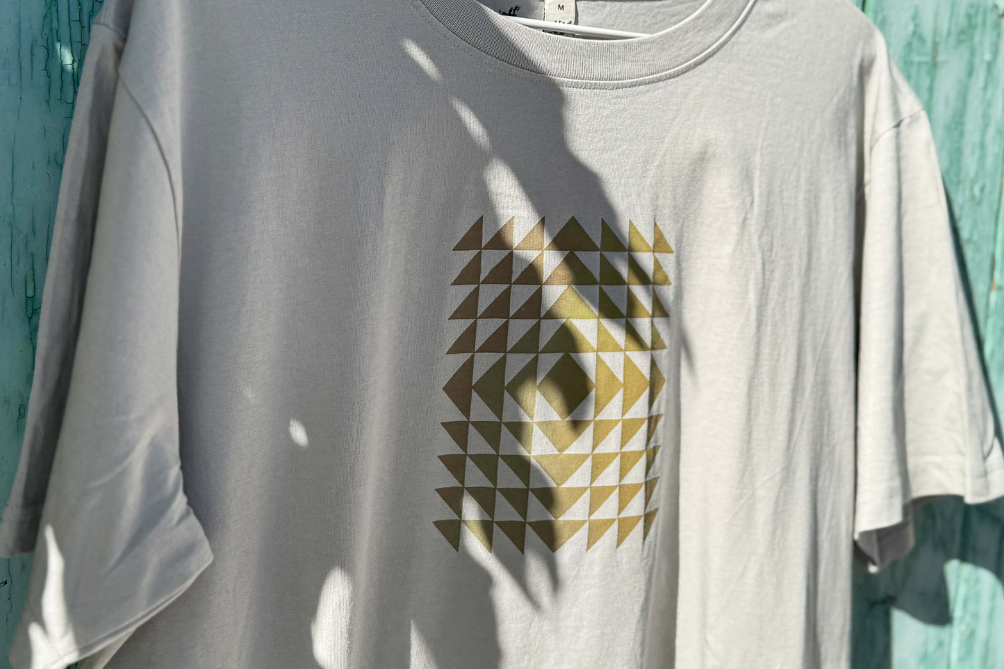 Detailansicht: Vorderseite eines T-Shirt in hellem grau vor blauen Hintergrund. Das Shirt zeigt ein minimalistisches und geometrisches Muster in der Farbe ocker auf der Brust.