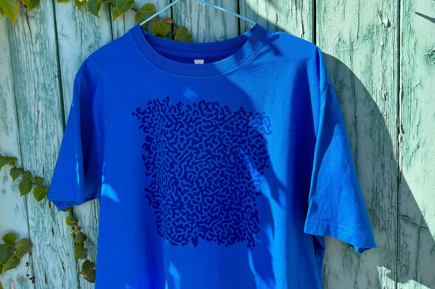 Detailansicht von einem blauen T-Shirt vor einem blauen Hintergrund. Das T-Shirt hat einen welligen Druck auf der Brust.