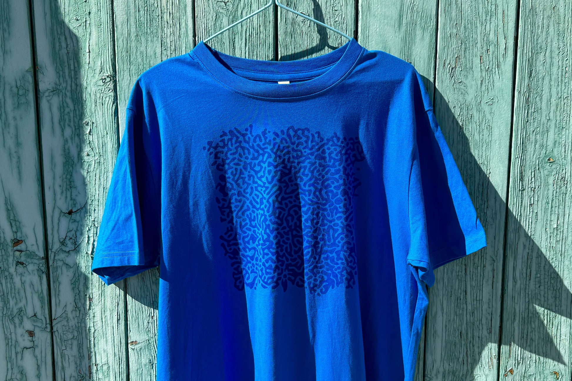 Detailansicht von einem blauen T-Shirt vor einem blauen Hintergrund. Das T-Shirt hat einen welligen Druck auf der Brust.