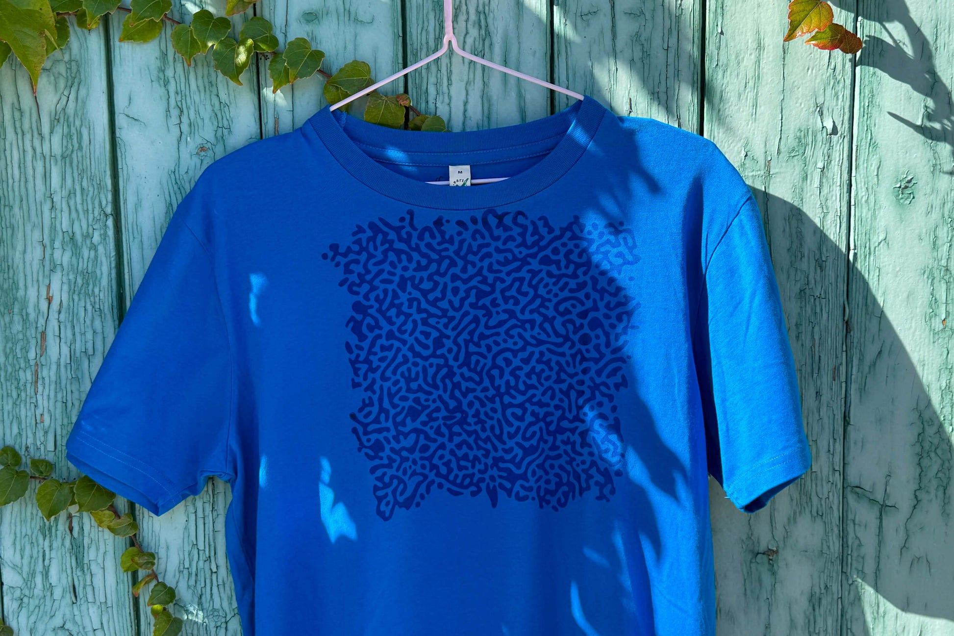 Detailansicht von einem blauen T-Shirt vor einem blauen Hintergrund. Das T-Shirt hat einen welligen Druck auf der Brust.