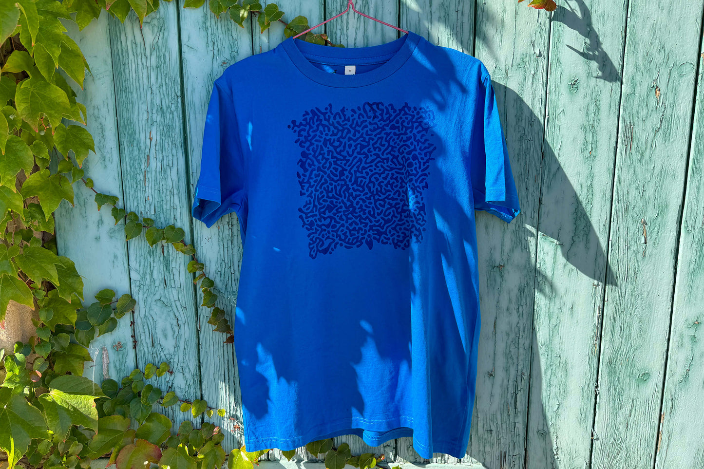 Gesamtansicht von einem blauen T-Shirt vor einem blauen Hintergrund. Das T-Shirt hat einen welligen Druck auf der Brust.