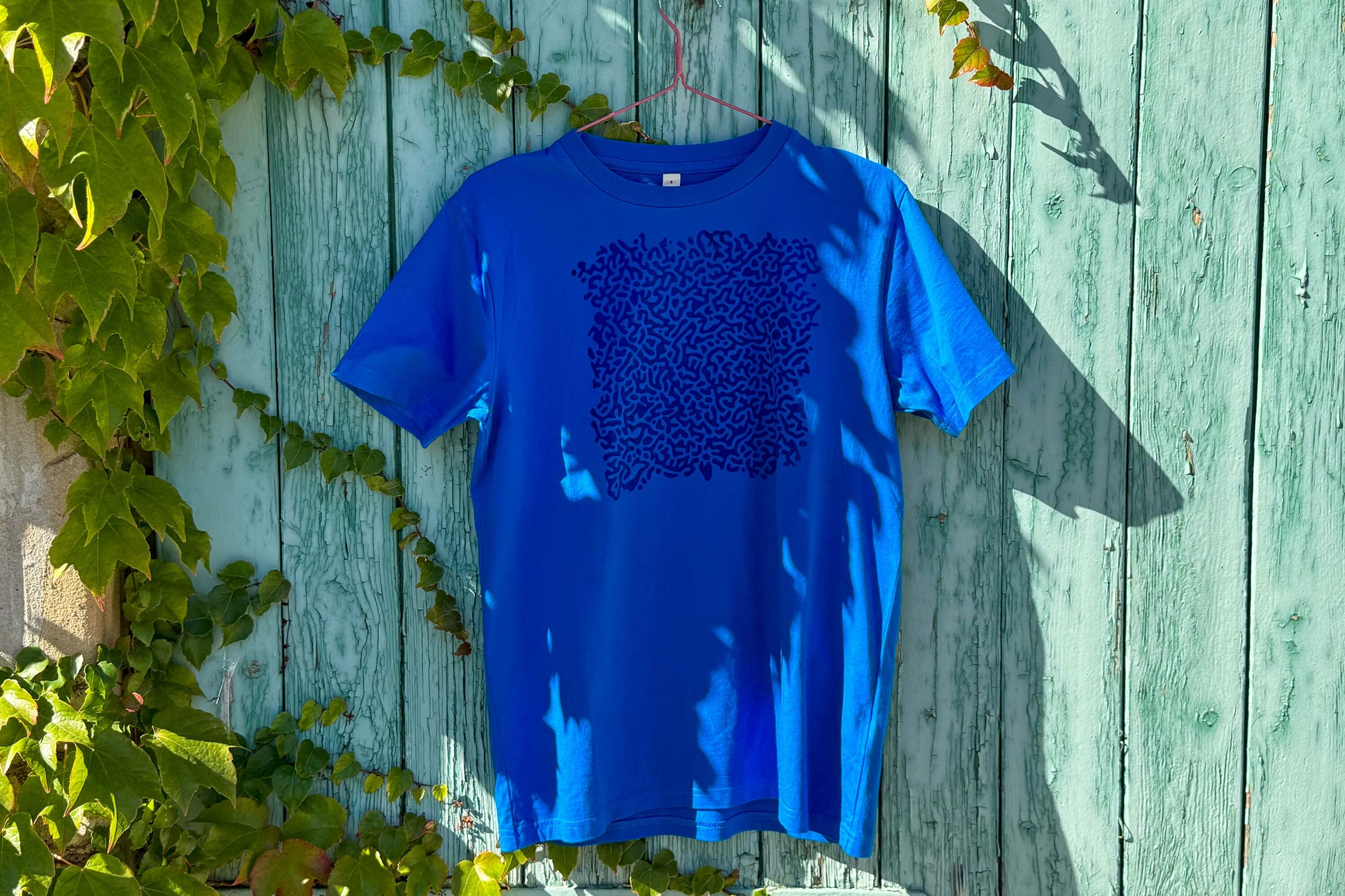 Gesamtansicht von einem blauen T-Shirt vor einem blauen Hintergrund. Das T-Shirt hat einen welligen Druck auf der Brust.