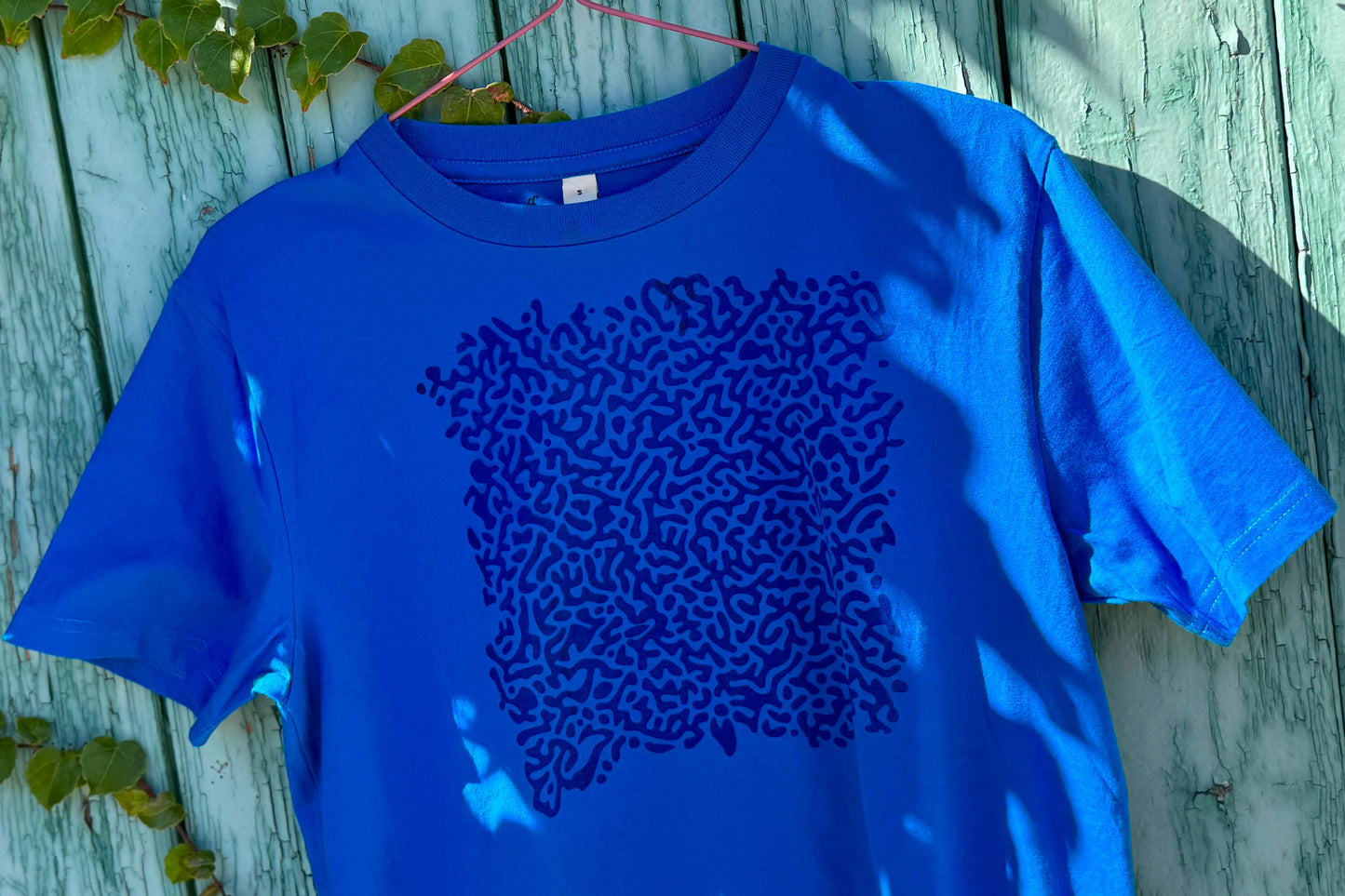 Detailansicht von einem blauen T-Shirt vor einem blauen Hintergrund. Das T-Shirt hat einen welligen Druck auf der Brust.