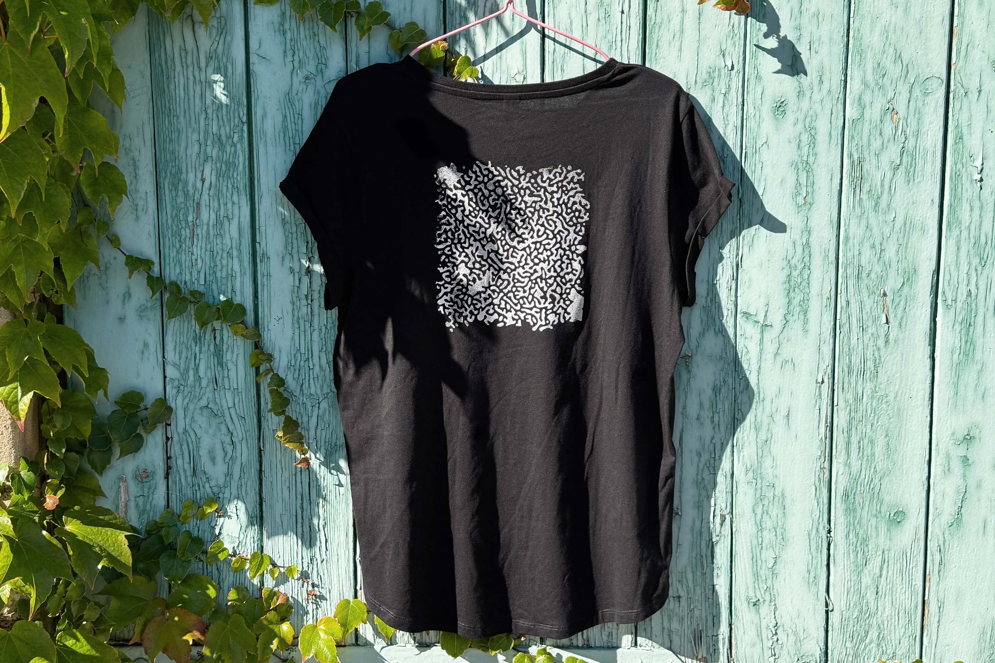 Hintere Gesamtansicht von einem schwarzen T-Shirt vor einem blauen Hintergrund. Das T-Shirt hat einen welligen Druck in silberner Farbe auf dem Rücken.