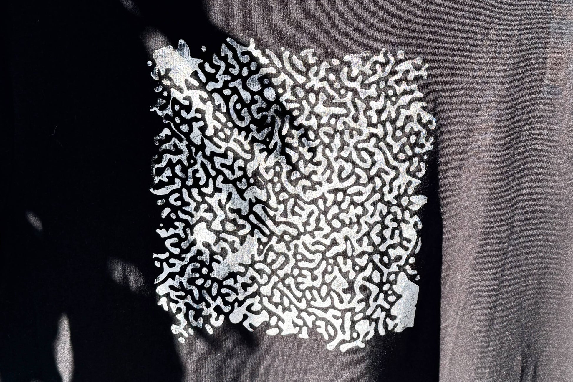 Hintere Detailansicht von einem schwarzen T-Shirt. Das T-Shirt hat einen welligen Druck in silberner Farbe auf dem Rücken.