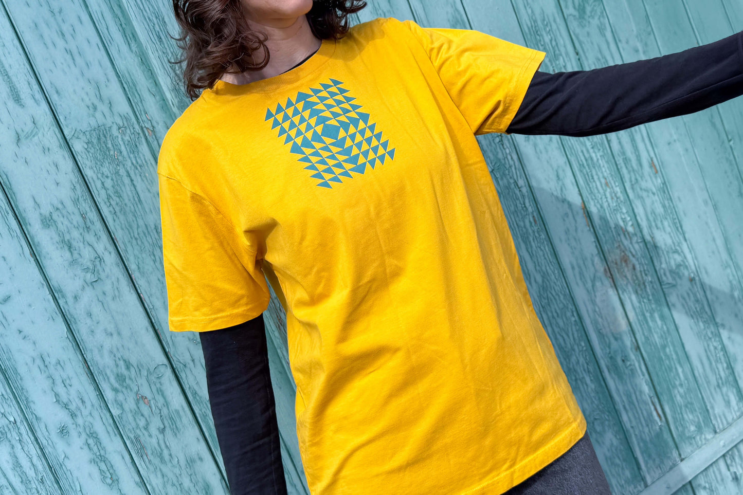 Gesamtansicht: Frau trägt ein T-Shirt in knalligem gelb vor blauen Hintergrund. Das Shirt zeigt ein minimalistisches und geometrisches Muster in der Farbe blau auf der Brust.