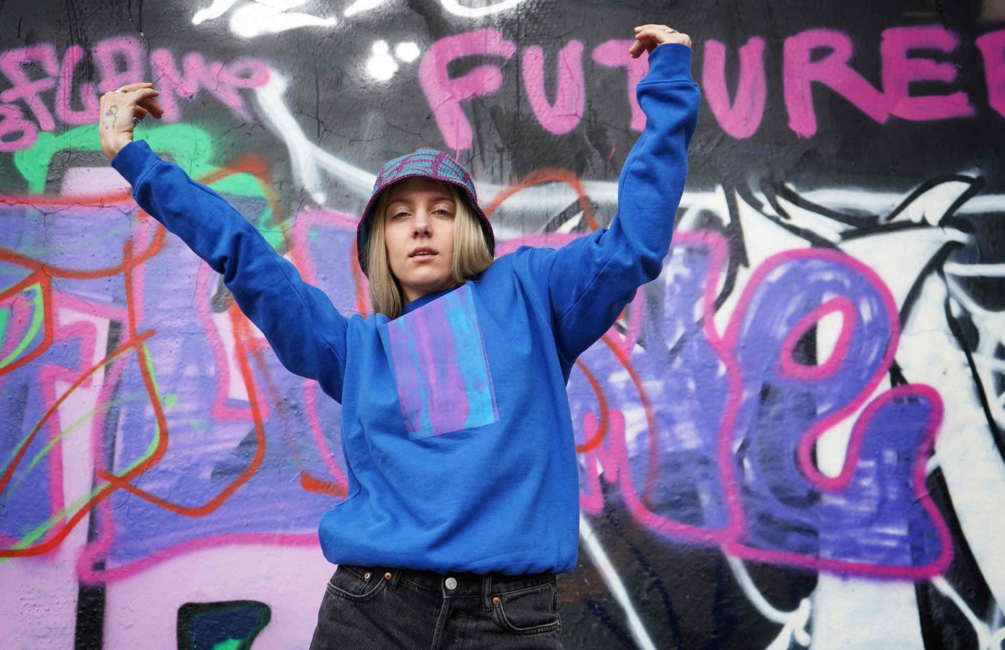 Frau steht vor Grafittiwand. Sie trägt Unisex Sweatshirt mit Siebdruck und von Hand gehäkeltem Anglerhut von jeff every piece a friend