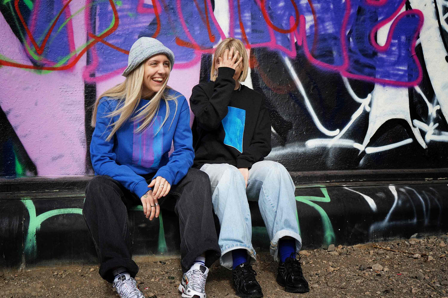 Zwei lachende Frauen vor Grafittiwand. Sie tragen Unisex Sweatshirts mit Siebdruck und von Hand gehäkelte Sommerhüte von jeff every piece a friend