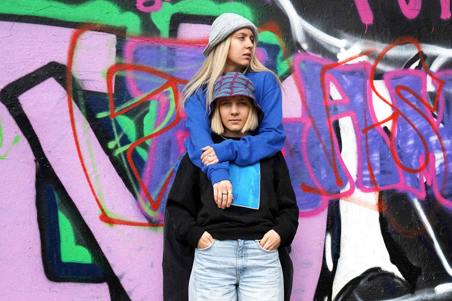 Zwei Frauen vor Grafittiwand. Sie tragen Unisex Sweatshirts mit Siebdruck und von Hand gehäkeltem Anglerhut von jeff every piece a friend