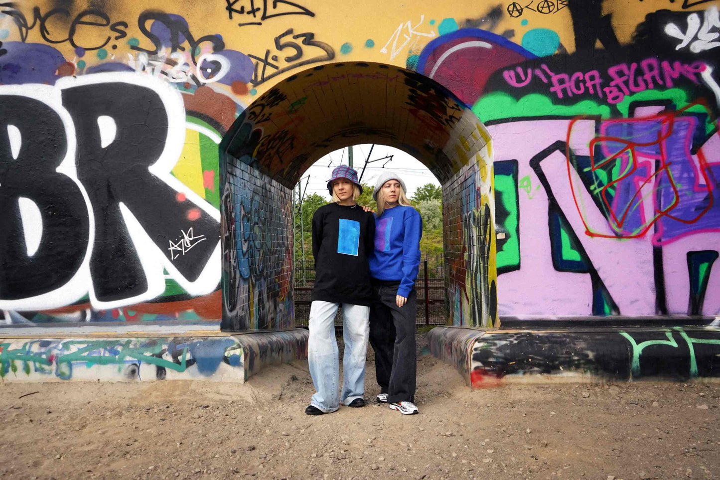 Zwei Frauen vor Grafittiwand. Sie tragen Unisex Sweatshirts mit Siebdruck und von Hand gehäkelte Sommerhüte von jeff every piece a friend