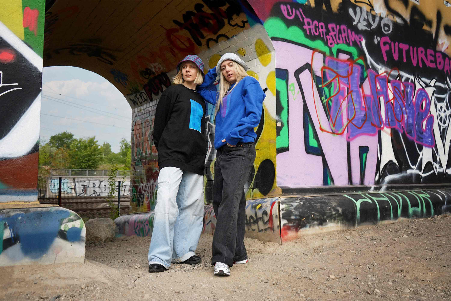 Zwei Frauen stehen vor Grafittiwand. Sie tragen Unisex Sweatshirts mit Siebdruck und von Hand gehäkelte Sommerhüte von jeff every piece a friend