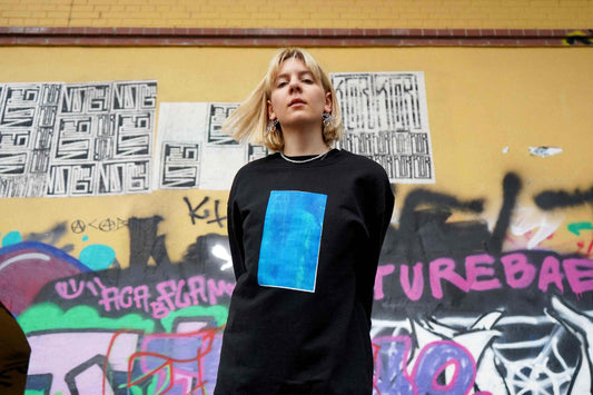 Junge Frau vor Grafittiwand. Sie trägt ein schwarzes Unisex Sweatshirt mit blauen Siebdruck von jeff every piece a friend