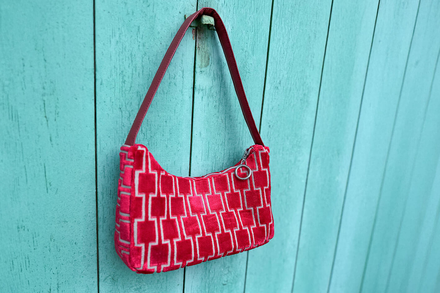 Gesamtansicht: Schultertasche im Vintagestil vor blauen Hintergrund. Die Tasche ist rot gemustert und aus Samt und hat einen silbernen Reißverschluss. Die Tasche wurde aus upcycled Materialien von Jeff Every Piece A Friend hergestellt.