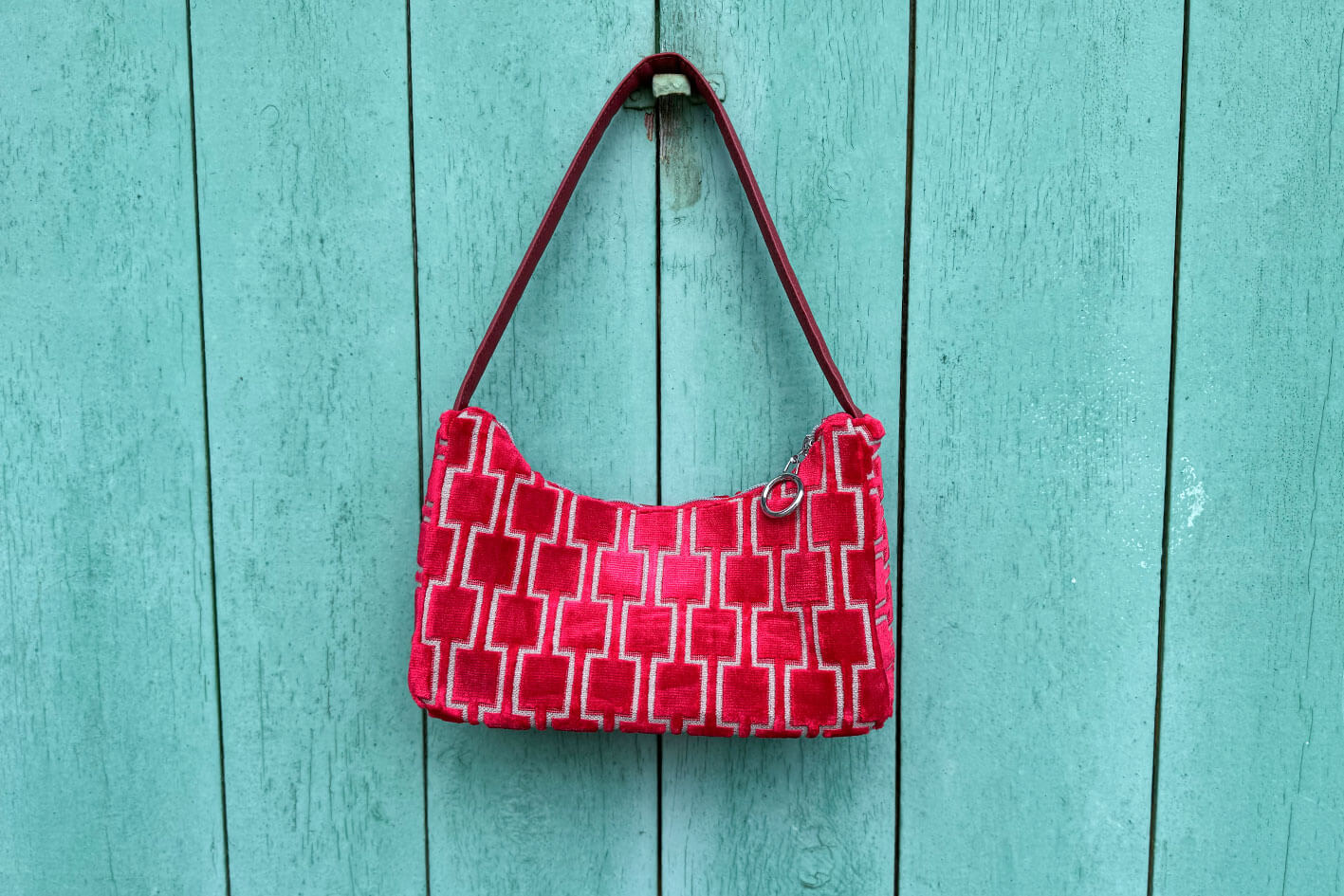 Gesamtansicht: Schultertasche im Vintagestil vor blauen Hintergrund. Die Tasche ist rot gemustert und aus Samt und hat einen silbernen Reißverschluss. Die Tasche wurde aus upcycled Materialien von Jeff Every Piece A Friend hergestellt.