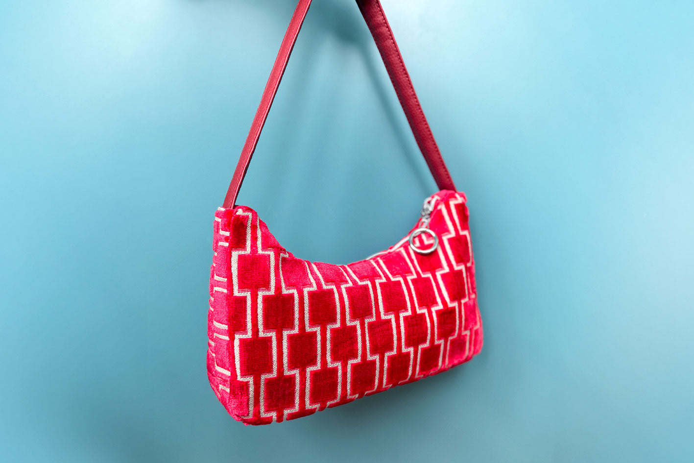 Gesamtansicht: Schultertasche im Vintagestil vor blauen Hintergrund. Die Tasche ist rot gemustert und aus Samt und hat einen silbernen Reißverschluss. Die Tasche wurde aus upcycled Materialien von Jeff Every Piece A Friend hergestellt.