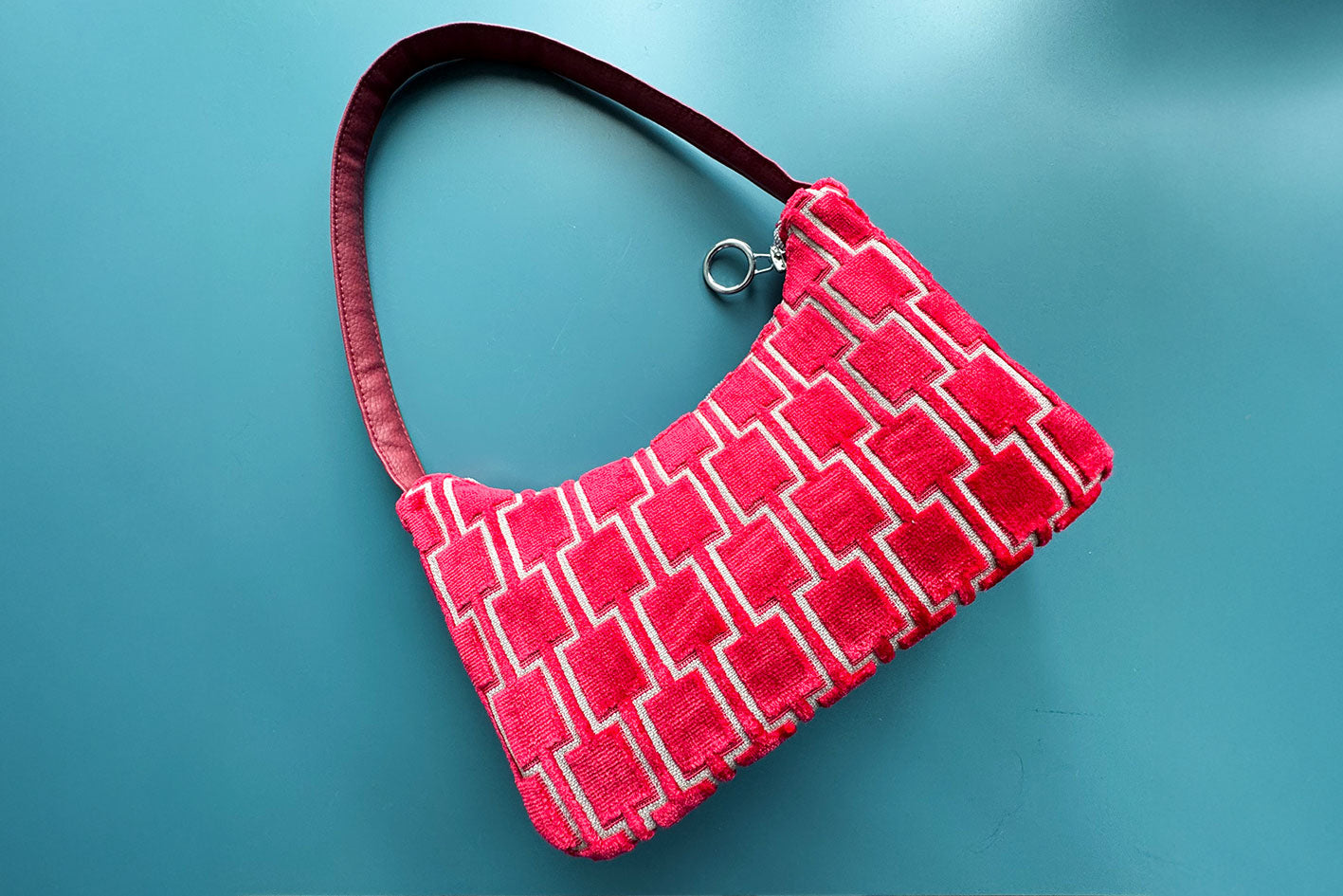 Gesamtansicht: Schultertasche im Vintagestil vor blauen Hintergrund. Die Tasche ist rot gemustert und aus Samt und hat einen silbernen Reißverschluss. Die Tasche wurde aus upcycled Materialien von Jeff Every Piece A Friend hergestellt.