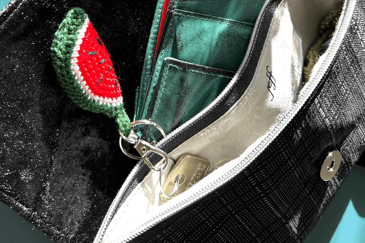 Close up der geöffneten umgehängten Clutch DANA hergestellt aus grau schwarzem Upcyling Polstermöbelstoff. Aus der Tasche ragt ein gehäkelter Wassermelonen-Schlüsselanhänger