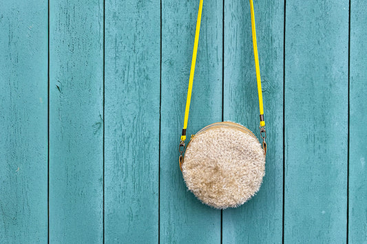 Runde Umhängetasche Peppa vor blauem Hintergrund. Tasche besteht aus upcycled Materialien mit gehäkeltem Deckel in der Farbe gold.