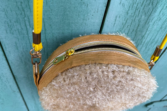 Detailansicht: Runde Umhängetasche Peppa vor blauem Hintergrund. Tasche besteht aus upcycled Materialien mit gehäkeltem Deckel in der Farbe gold.