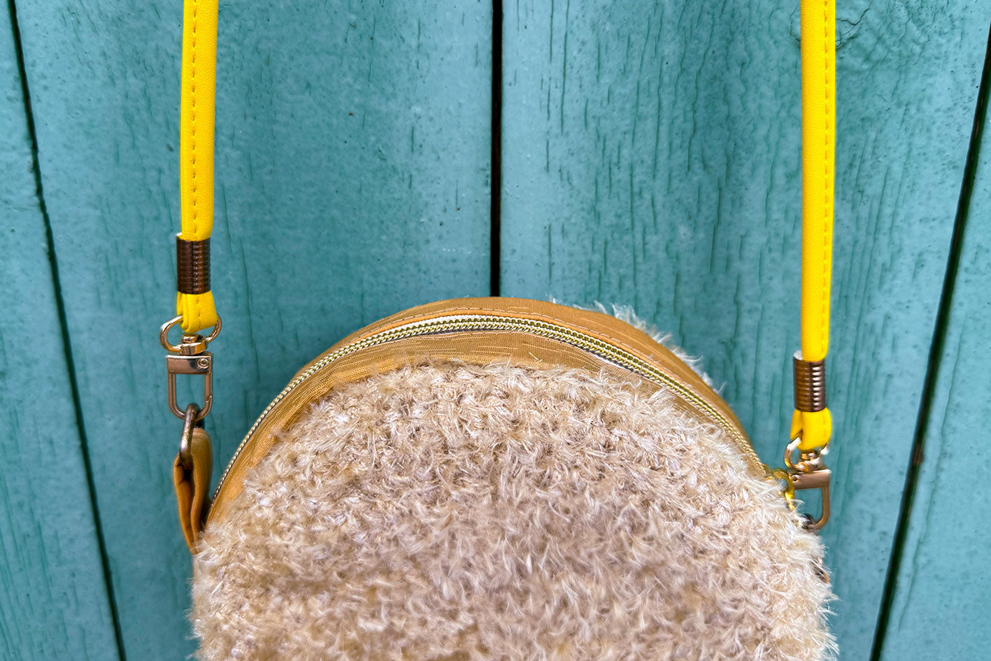 Detailansicht Reißverschluss: Runde Umhängetasche Peppa vor blauem Hintergrund. Tasche besteht aus upcycled Materialien mit gehäkeltem Deckel in der Farbe gold.