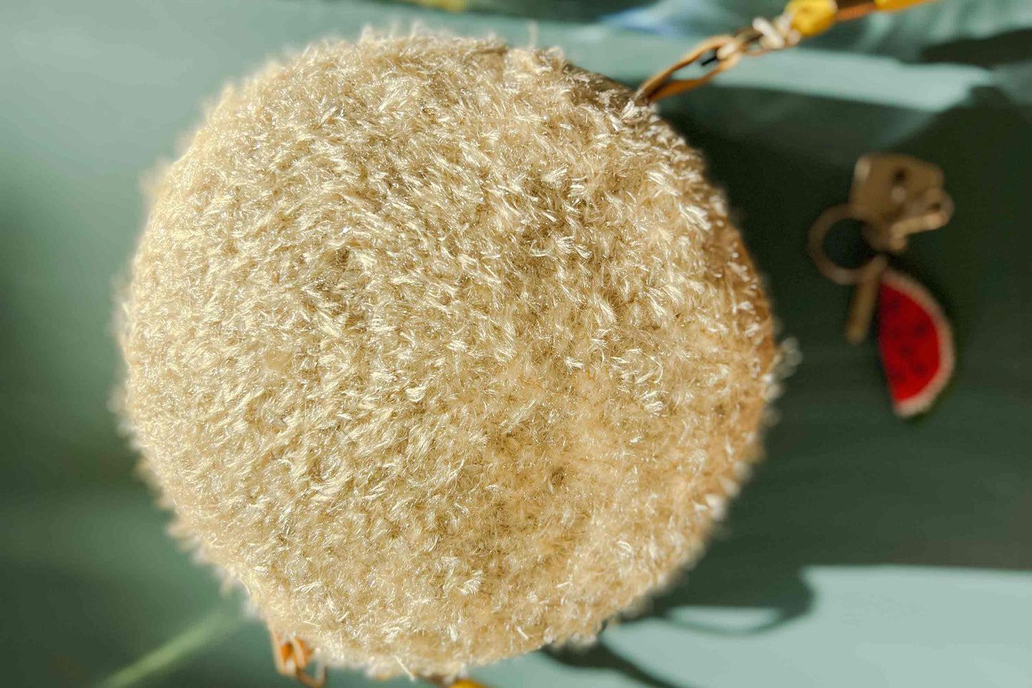 Detailansicht des goldenen, gehäkelten Deckels der runden Umhängetasche Peppa vor blauem Hintergrund. Tasche besteht aus upcycled Materialien. Im Hintergrund liegt ein Schlüssel mit einem Anhänger.