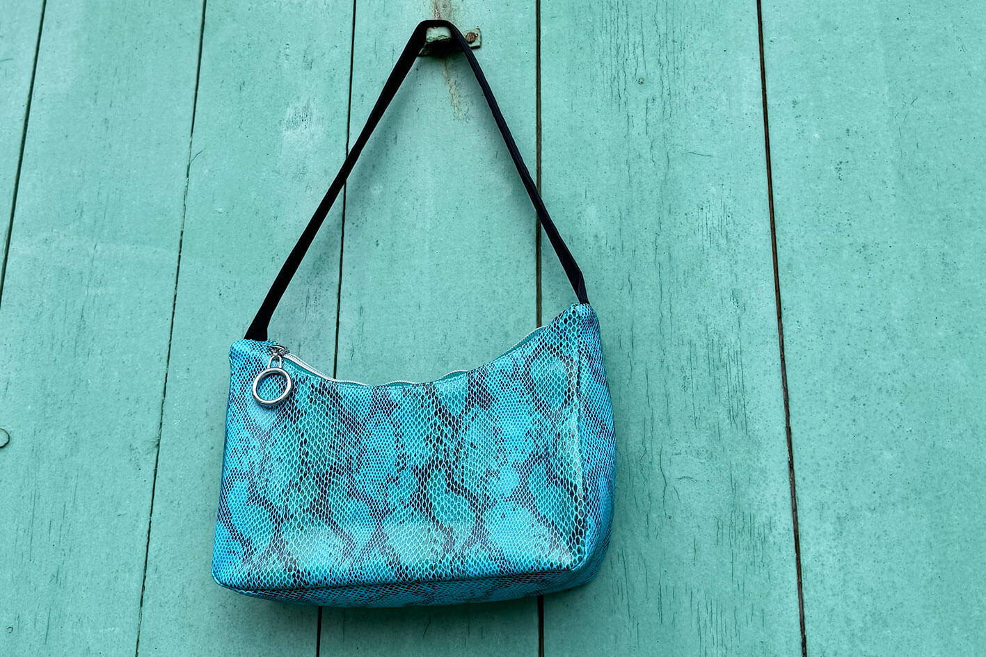 Gesamtansicht blaue Schultertasche in Schlangeleder Optik vor blauem Hintergrund mit schwarzem Gurtband. Hergestellt aus upcycled Materialien von Jeff Every Piece A Friend.