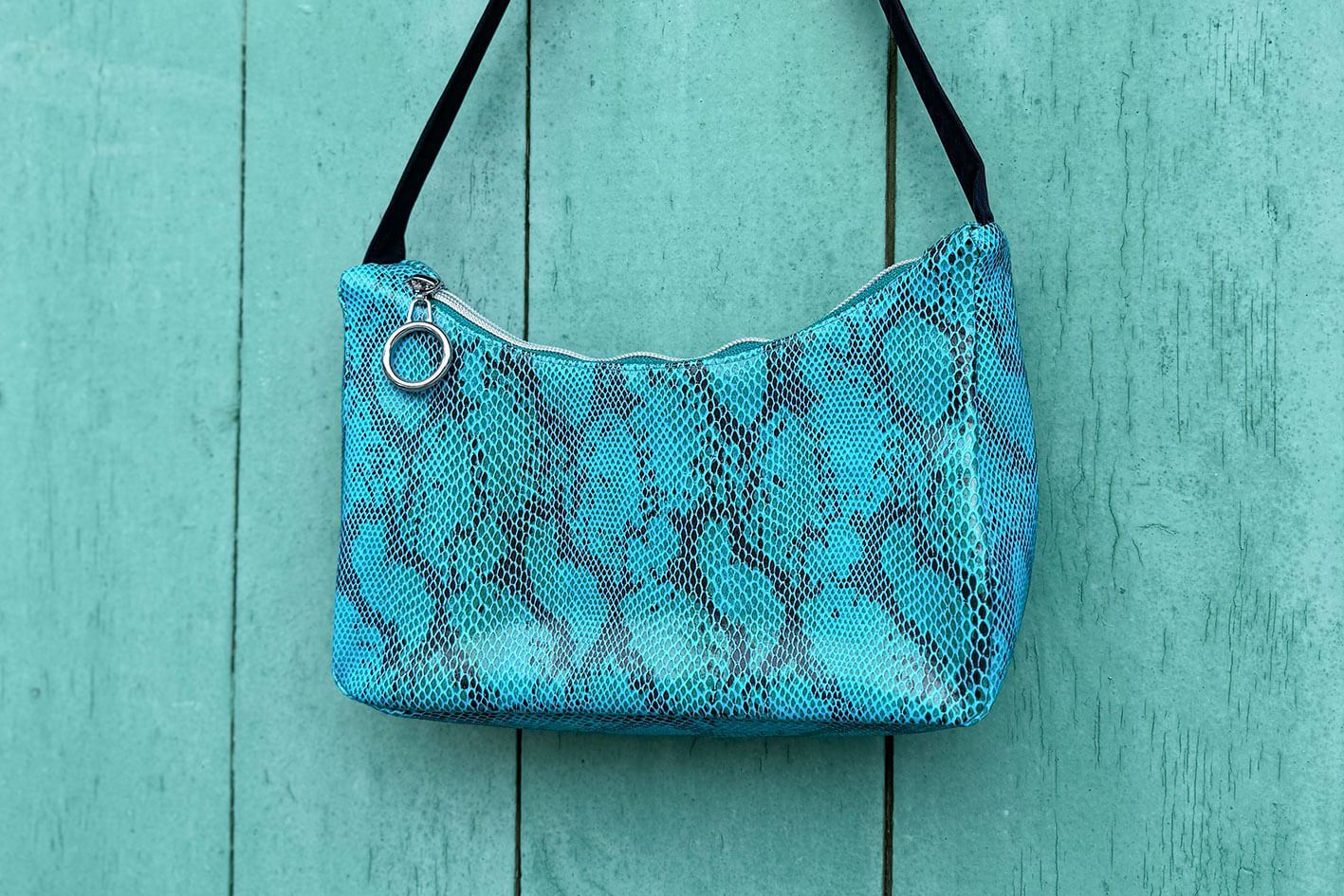 Gesamtansicht blaue Schultertasche in Schlangeleder Optik vor blauem Hintergrund mit schwarzem Gurtband. Hergestellt aus upcycled Materialien von Jeff Every Piece A Friend.