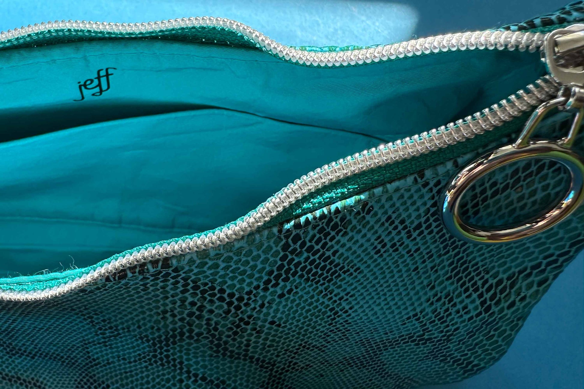 Detailansicht von Innenfach. Blaue Schultertasche in Schlangeleder Optik vor blauem Hintergrund mit schwarzem Gurtband und silbernen Reißverschluss. Hergestellt aus upcycled Materialien von Jeff Every Piece A Friend.
