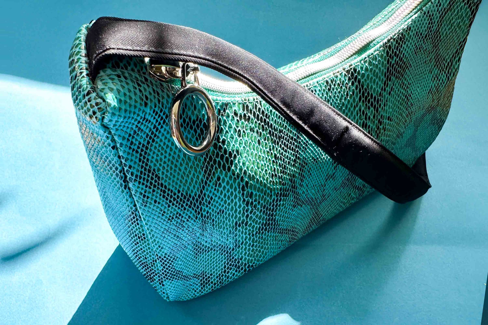 Detailansicht blaue Schultertasche in Schlangeleder Optik vor blauem Hintergrund mit schwarzem Gurtband und silbernen Reißverschluss. Hergestellt aus upcycled Materialien von Jeff Every Piece A Friend.