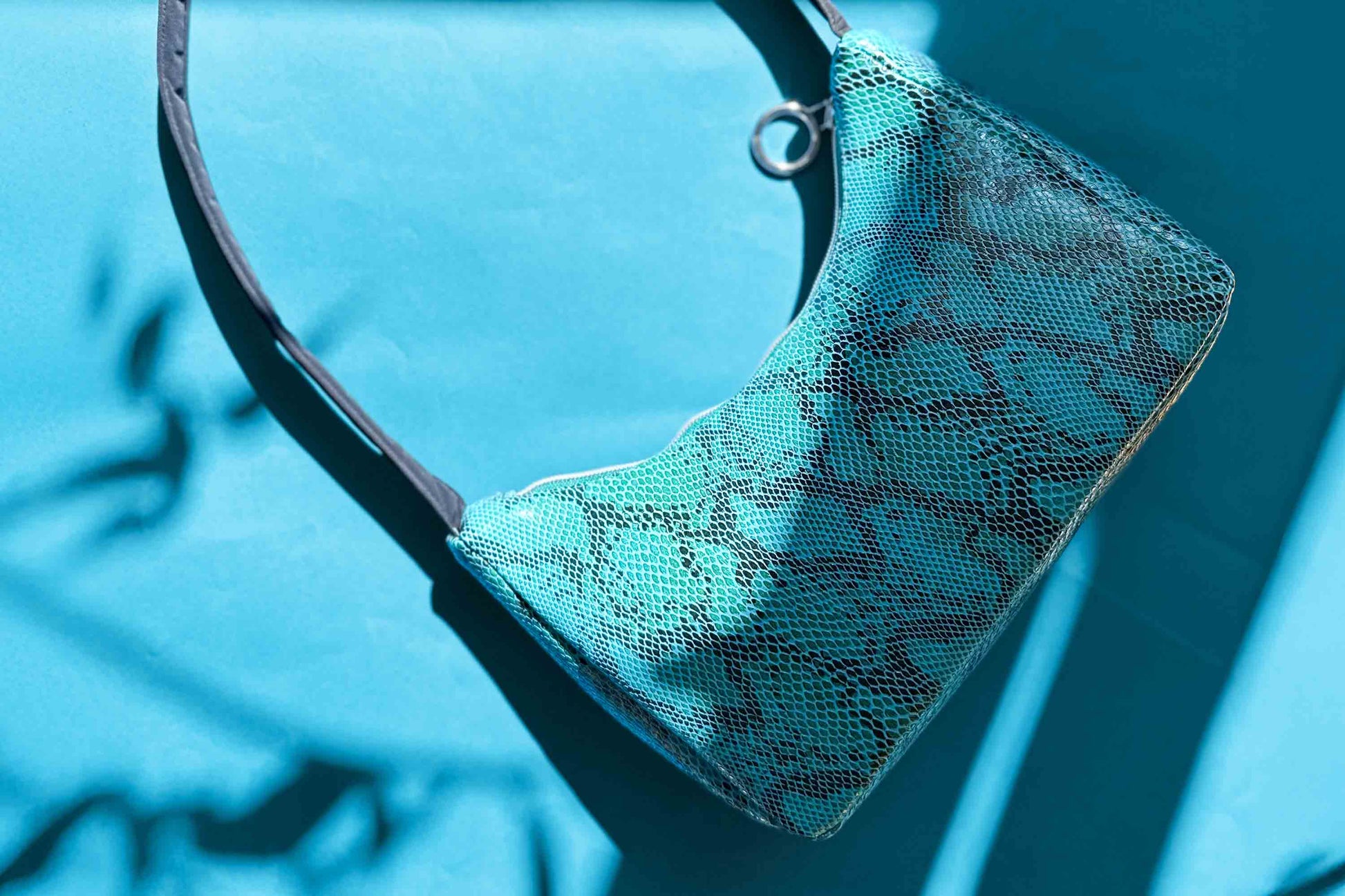 Gesamtansicht blaue Schultertasche in Schlangeleder Optik vor blauem Hintergrund mit schwarzem Gurtband. Hergestellt aus upcycled Materialien von Jeff Every Piece A Friend.
