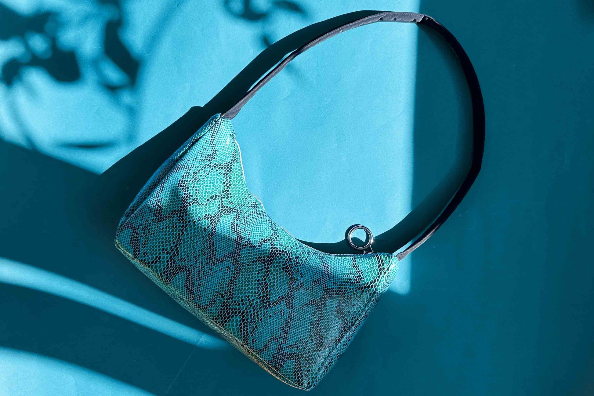 Gesamtansicht blaue Schultertasche in Schlangeleder Optik vor blauem Hintergrund mit schwarzem Gurtband. Hergestellt aus upcycled Materialien von Jeff Every Piece A Friend.