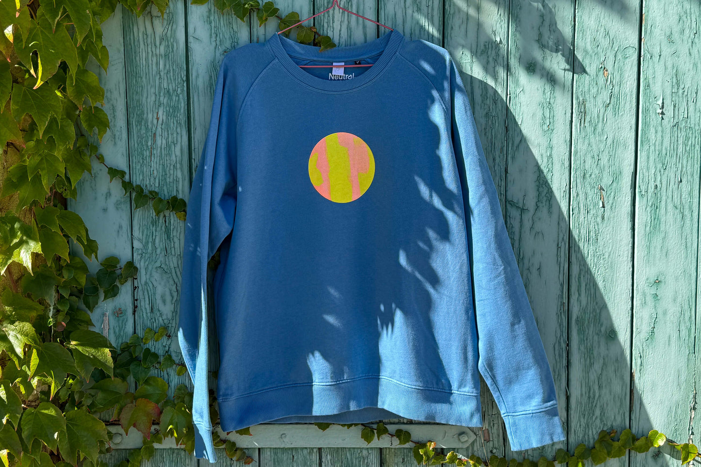 Vorderansicht von blauem Sweatshirt vor blauem Hintergrund. Der Pulli hat einen aufgedruckten Kreis in gelb-orange.