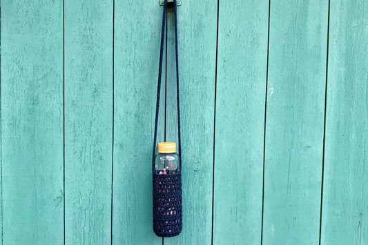 Gehäkelter, dunkelblauer Flaschenhalter CHRISTA von Jeff Every Piece A Friend vor blauem Hintergrund.