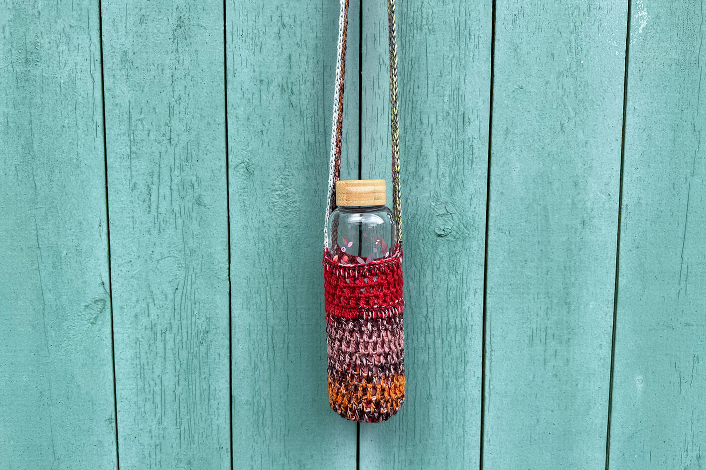 Gehäkelter, rot braun oranger Flaschenhalter CHRISTA von Jeff Every Piece A Friend vor blauem Hintergrund.