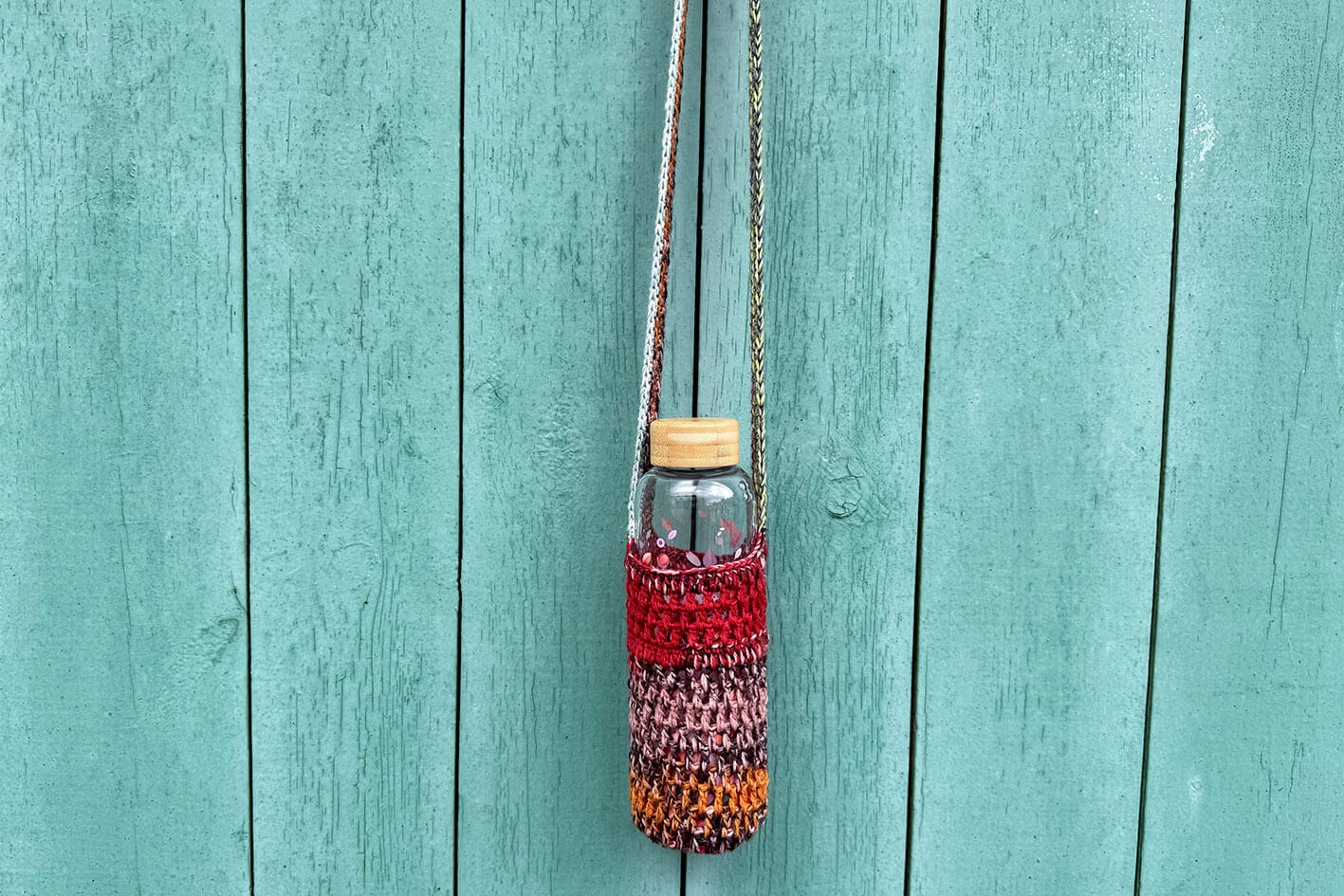 Gehäkelter, rot braun oranger Flaschenhalter CHRISTA von Jeff Every Piece A Friend vor blauem Hintergrund.