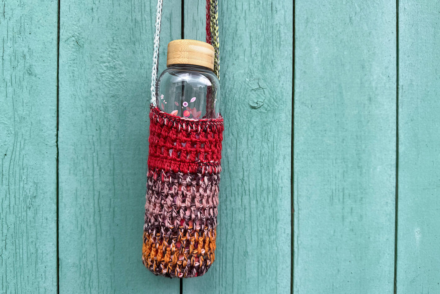 Gehäkelter, rot braun oranger Flaschenhalter CHRISTA von Jeff Every Piece A Friend vor blauem Hintergrund.
